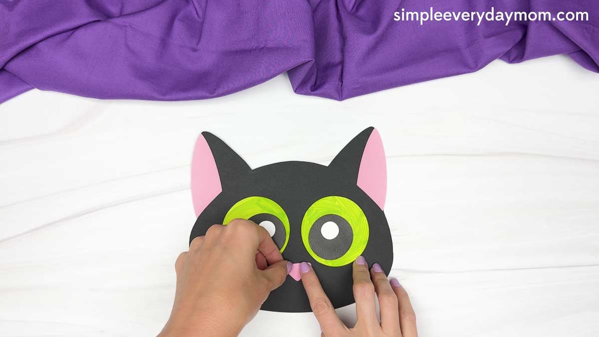 Hands place a small pink triangular nose onto the black cat face with green eyes and pink inner ears.