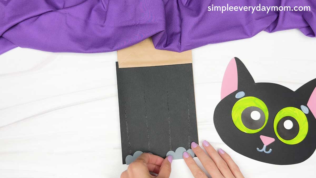 Hands attach gray paper paws to the bottom of the black paper bag body while the finished cat head rests beside it.