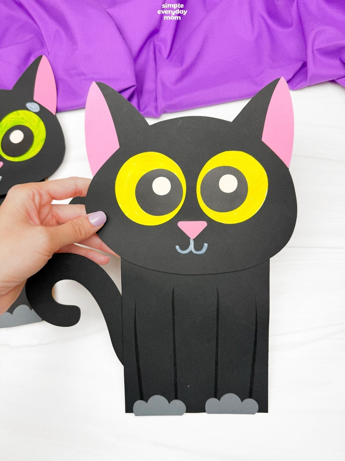 A hand holds a finished black paper cat puppet with large yellow eyes, pink ears and nose, gray paws, and a curled tail; part of a green-eyed cat puppet appears in the background.