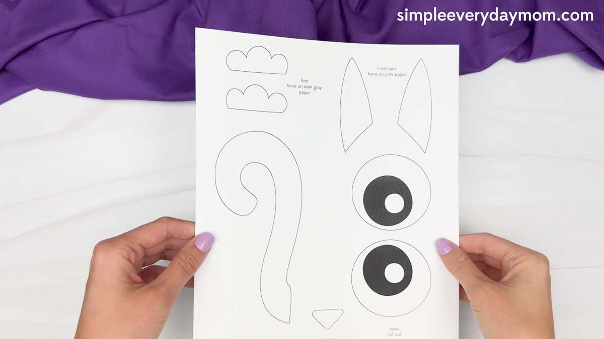 Hands hold a printable black cat craft template featuring outlines for the tail, eyes, inner ears, nose, and paws.