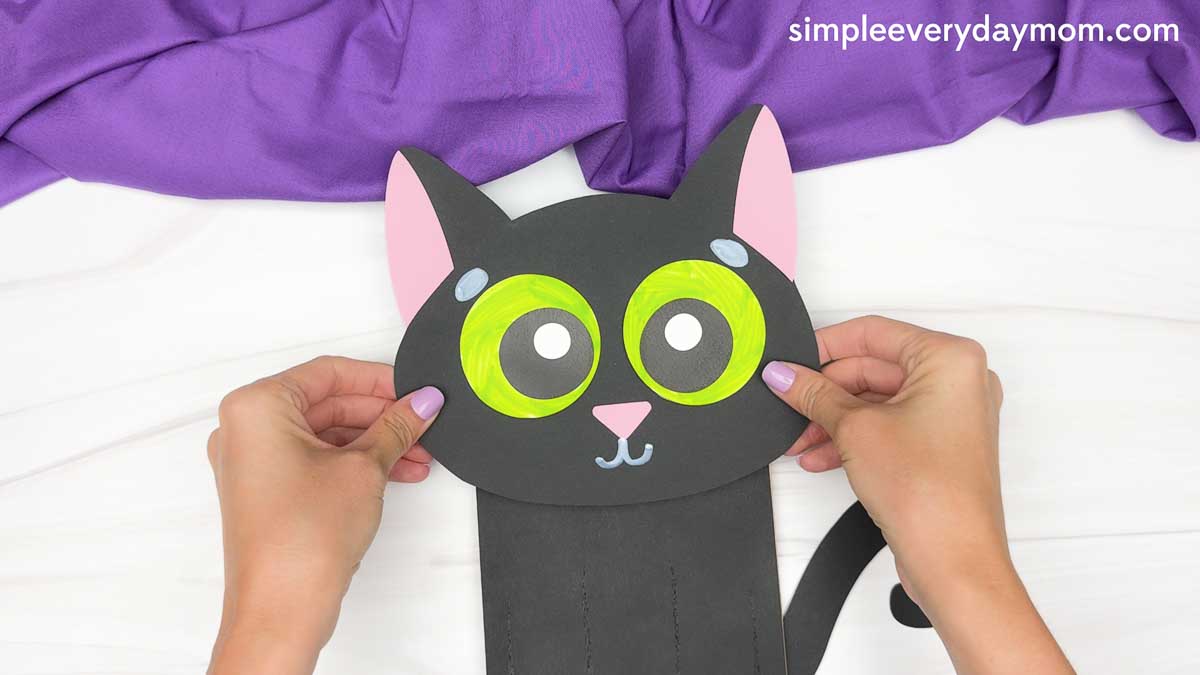 Hands position the completed black cat head onto the paper bag body, with green eyes, pink ears, gray details, and a curled tail.