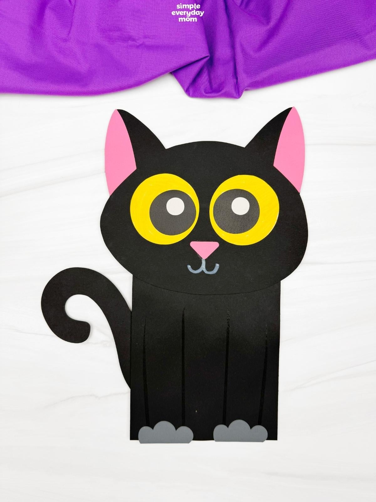 Finished black paper cat puppet with large yellow eyes, pink ears and nose, gray paws, and a curled tail on a white background.