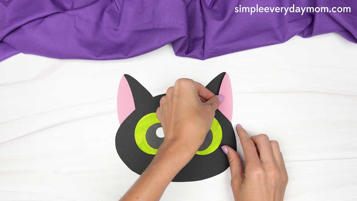Hands attach pink inner-ear pieces to the pointed ears of a black cat head cutout.