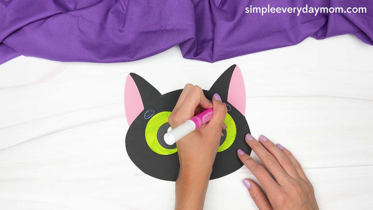 A hand uses a marker to add small gray details above the green eyes of the black cat face.
