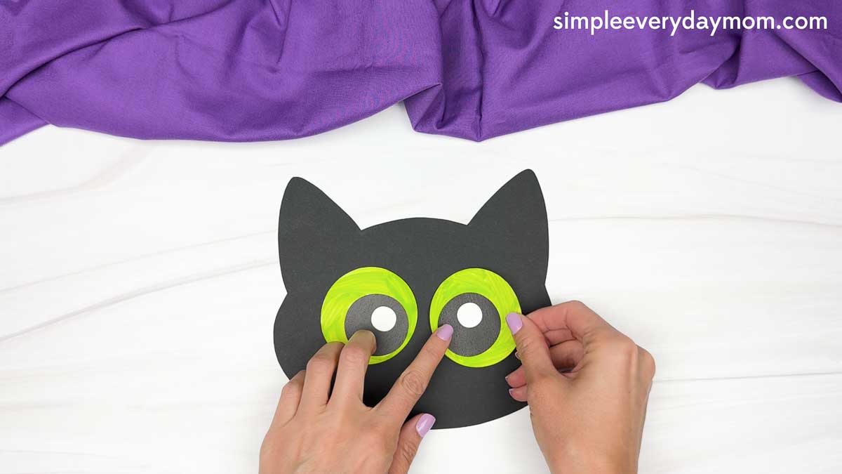 Hands attach large green paper eyes to a black cat head cutout.