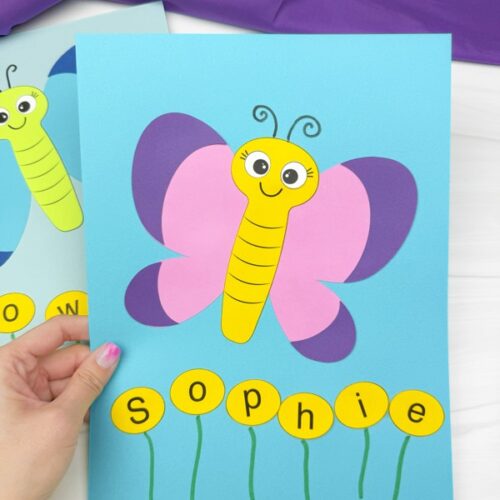 A hand holds a completed butterfly name craft featuring a smiling butterfly with pink and purple wings on a blue background. Below, yellow circles spell "Sophie," with green marker-drawn stems resembling flowers. Another similar craft with the name "Rowan" is partially visible in the background.