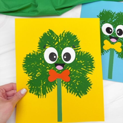 A hand holding a fork-painted shamrock craft with cartoon eyes, a smiling mouth, and a red bow tie on a yellow background. Another similar craft with a blue background is partially visible in the background.