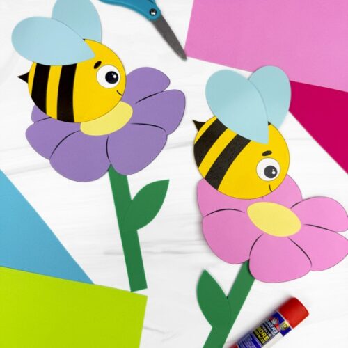 Two colorful paper bumblebee crafts on purple and pink flowers, surrounded by crafting supplies including scissors, glue stick, and sheets of colored paper on a white surface.