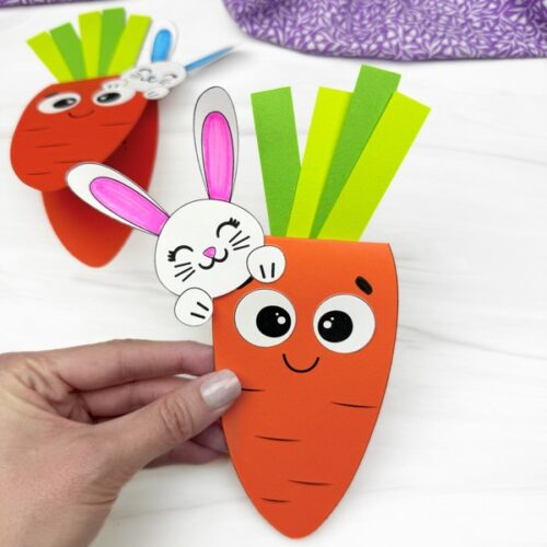 A handmade paper craft of a smiling carrot with green paper leaves and a cheerful bunny with pink ears peeking from behind is being held by a person's hand. Another similar carrot craft is slightly blurred in the background on a white surface with a purple patterned fabric.