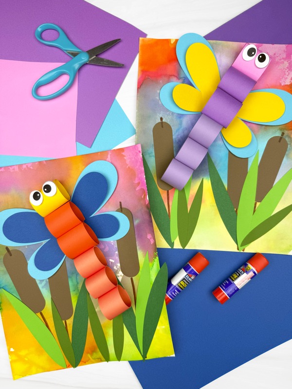 Two colorful 3D dragonfly crafts made with paper chains and bright wings, set on watercolor backgrounds with cattails and green grass. Surrounded by craft supplies like scissors, glue sticks, and construction paper.