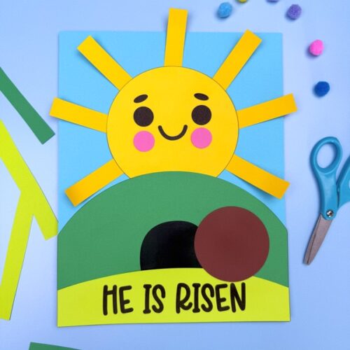 A "He Is Risen" Easter craft for kids featuring a cheerful yellow sun with a smiling face, a brown hill with a black tomb entrance, and a movable brown stone. The craft is made from colorful paper and is set against a blue background with scattered crafting supplies.