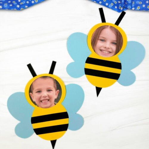 A children's craft featuring two paper honey bee cutouts with real children's photos as their faces. The bees have yellow bodies with black stripes, light blue wings, black antennae, and small black triangles as stingers, set against a white background with a blue floral fabric partially visible at the top.