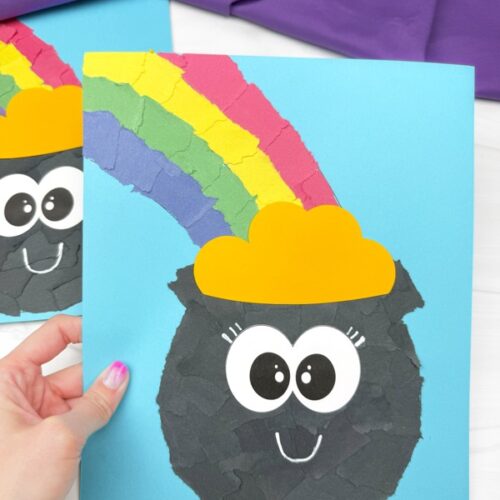 A hand holds up one of the pot of gold crafts, showing its torn paper texture, large cartoon eyes, and curved smile.