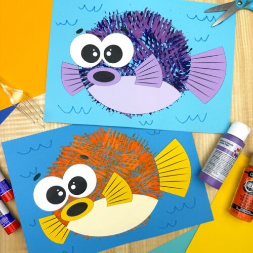 Two colorful fork-painted puffer fish crafts—one purple and one orange—displayed on a wooden surface with paint, glue sticks, scissors, and colored paper nearby.