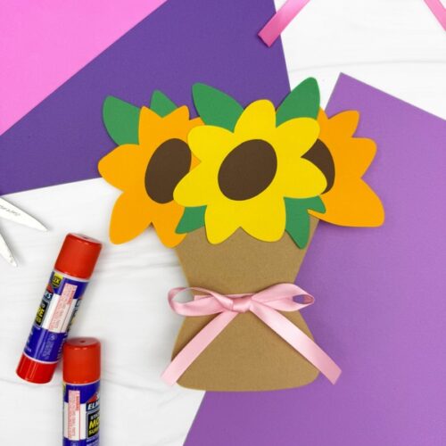 Finished paper sunflower bouquet craft with three sunflowers in a brown paper pot, tied with a pink ribbon, displayed on purple and pink construction paper with glue sticks nearby.