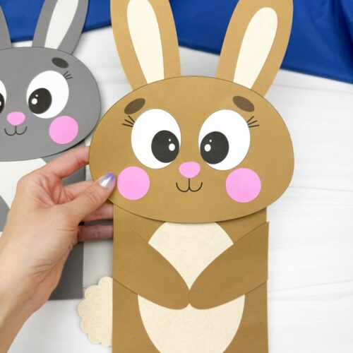 A person is holding a brown paper bag bunny puppet with large eyes, pink cheeks and nose, folded arms, cream-colored belly, and cotton tail; a gray bunny puppet is partially visible in the background on a white surface with a blue fabric.
