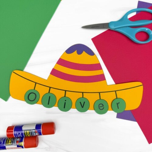Colorful paper sombrero craft labeled “Oliver” with green letter circles, surrounded by scissors, glue sticks, and bright construction paper.