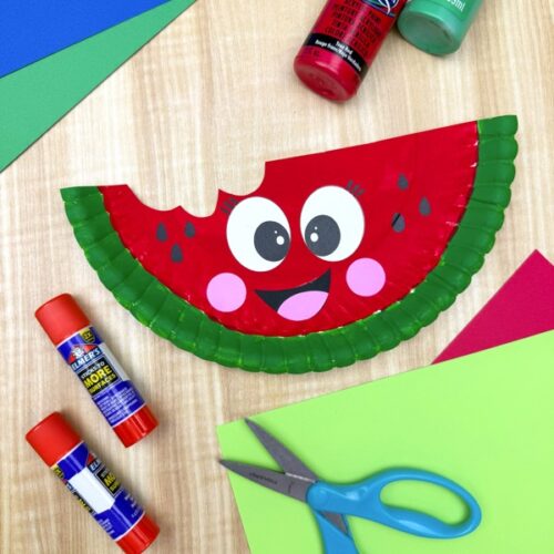 Smiling watermelon paper plate craft with a bite mark, surrounded by red and green paint bottles, glue sticks, scissors, and colorful paper. A bright and playful kids’ craft setup perfect for summer.