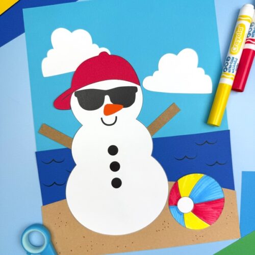 A completed beach snowman craft is set on a blue surface, decorated with a red cap, sunglasses, clouds, waves, sand, and a colorful beach ball, surrounded by scissors, markers, and colored paper.