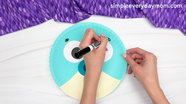 A person uses a black marker to draw eyebrows above two large cartoon eyes on a teal and cream-colored paper plate bird craft with a blue beak, placed on a white surface with a purple patterned cloth in the background.