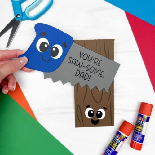 Father’s Day saw card craft made from construction paper, featuring a smiling blue saw and wood piece with the message “You’re SAW-some, Dad!” A simple and fun DIY project for preschoolers using scissors, glue sticks, and a free printable template.