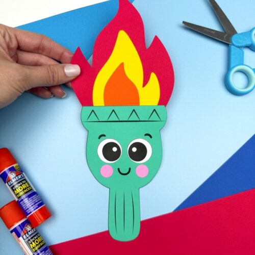 A hand holds up a completed Statue of Liberty torch craft with a happy face and colorful paper flame, set against a bright blue and red background with glue sticks, scissors, and craft paper nearby.