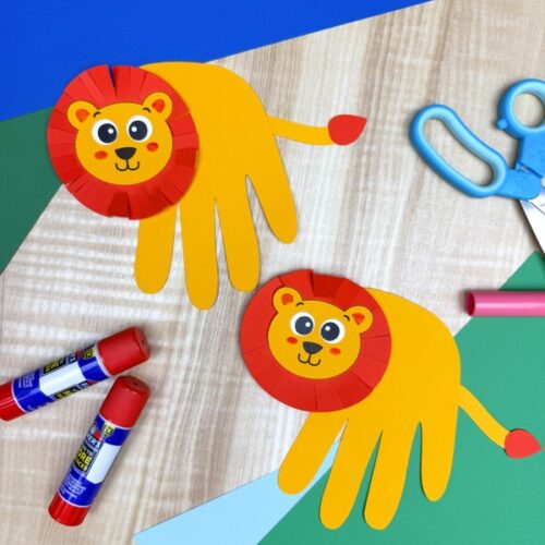 Two completed handprint lion crafts are displayed on a wooden surface surrounded by glue sticks, scissors, and colored paper, showing a finished result with a circular mane, hand-shaped body, and tail tip—all made from paper.