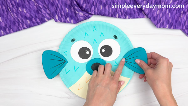 A person attaches blue paper wings to the sides of a bird craft made from a painted paper plate, which features large eyes, a blue beak, drawn-on feather patterns, and a two-tone design with teal on top and cream on the bottom.