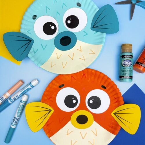 Two colorful paper plate pufferfish crafts are displayed on a blue background, one teal with dark teal fins and beak, and the other orange with yellow fins and beak, each featuring large eyes and hand-drawn scale patterns. Craft supplies like markers, scissors, and paint bottles surround the fish.