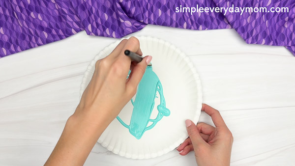 Hands painting a white paper plate with teal paint using a paintbrush, with a purple patterned fabric in the background.