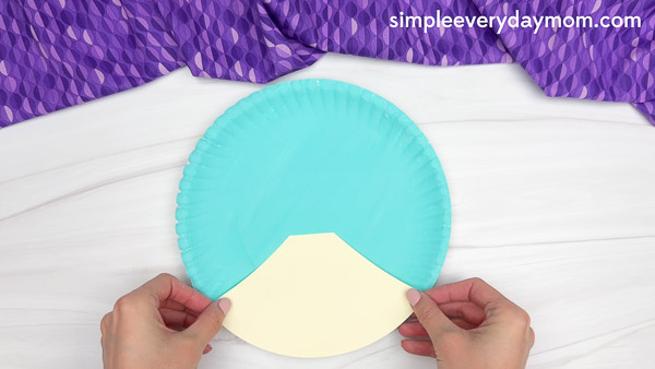 Hands holding a partially completed craft made from a paper plate painted teal, with a cream-colored semi-circle piece glued to the bottom center, against a white surface and purple patterned fabric background.