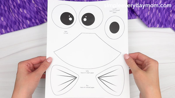 Hands holding a black and white printable frog craft template that includes large cartoon eyes, a mouth, and labeled body parts to cut out and trace on colored paper, against a white background with a purple patterned fabric.