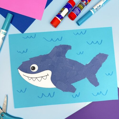 Torn paper shark craft surrounded by glue sticks, scissors, and markers on colorful paper. Easy ocean-themed art project for preschoolers and kindergarten kids.