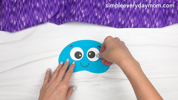 Hands are placing a large paper eye onto a blue jellyfish cutout during an ocean-themed kids craft activity.