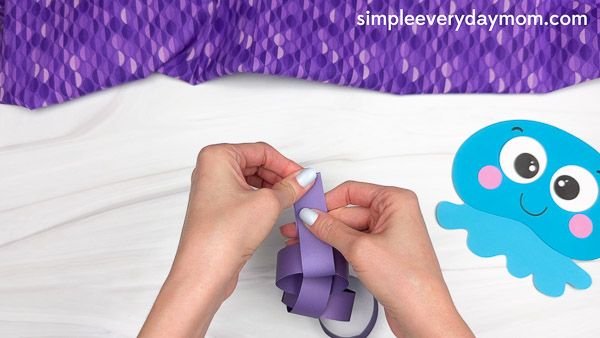 Hands are linking a purple paper strip into a chain to form the jellyfish's tentacles for an ocean-themed kids craft project.