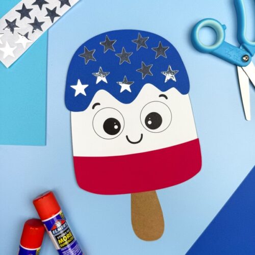 A completed 4th of July popsicle craft made from paper is displayed on a blue background, decorated with silver and white star stickers, next to glue sticks, scissors, and colored paper.