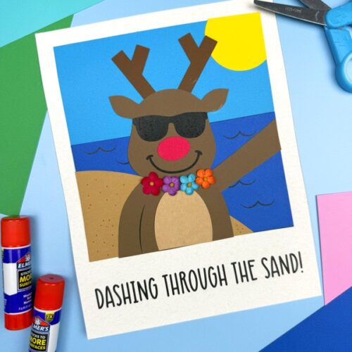 A completed reindeer beach craft is shown on a blue surface, featuring a reindeer with sunglasses, a red nose, and a colorful flower lei, standing on a sunny beach with the caption “DASHING THROUGH THE SAND!” below the scene.