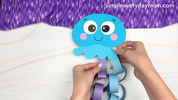 Hands are attaching the finished paper chain tentacles to the bottom of a blue jellyfish cutout as part of an ocean-themed kids craft.