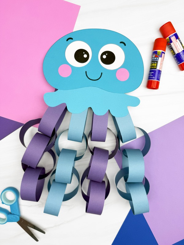 A completed blue paper chain jellyfish craft is displayed on a colorful background with glue sticks and scissors nearby, featuring a smiling face and tentacles made from alternating purple and blue paper loops—ideal for an ocean-themed kids activity.