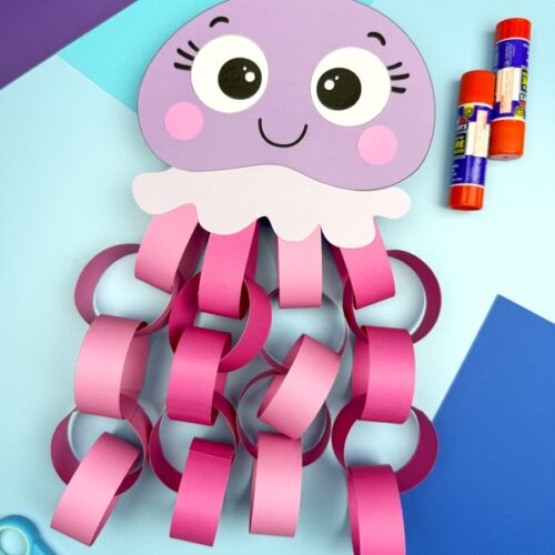 A completed purple jellyfish paper chain craft is displayed on a colorful background, featuring a smiling face and pink looped tentacles, with scissors, glue sticks, and craft paper nearby—perfect for an ocean-themed craft for kids.