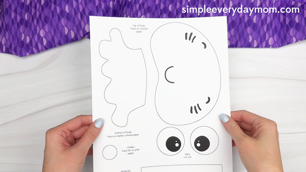Hands are holding up a printed jellyfish craft template sheet that includes outlines for the body, eyes, cheeks, and mouth—used to create an ocean-themed paper chain jellyfish craft for kids.