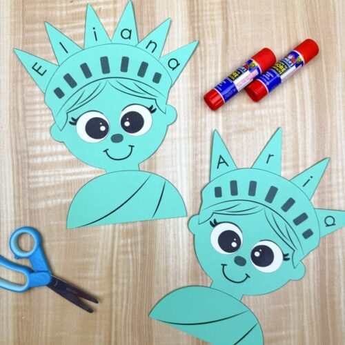 Two completed green paper Statue of Liberty crafts labeled “Eliana” and “Aria” with large cartoon eyes and smiling faces, displayed on a light wood surface with blue scissors and two red glue sticks nearby for a kids’ craft activity.