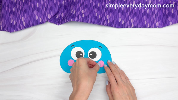 Hands are pressing pink paper cheeks onto a blue jellyfish head cutout as part of assembling an ocean-themed kids craft.