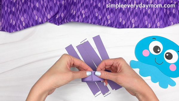 Hands are forming a loop with a purple paper strip to create a chain link, preparing the tentacles for a blue jellyfish paper craft as part of an ocean-themed kids activity.