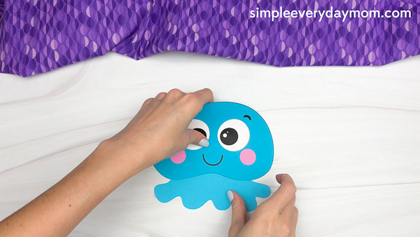 Hands are attaching the second eye to a blue jellyfish cutout, assembling the face as part of an ocean-themed kids paper craft.