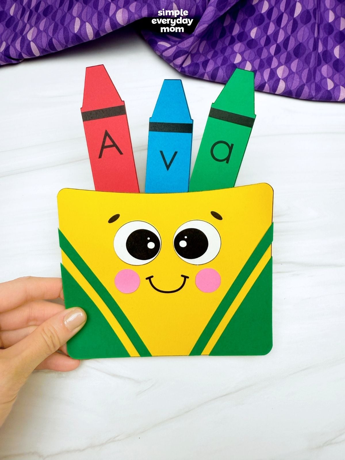 Hand holding a finished crayon box name craft labeled “Ava” with three colored paper crayons.