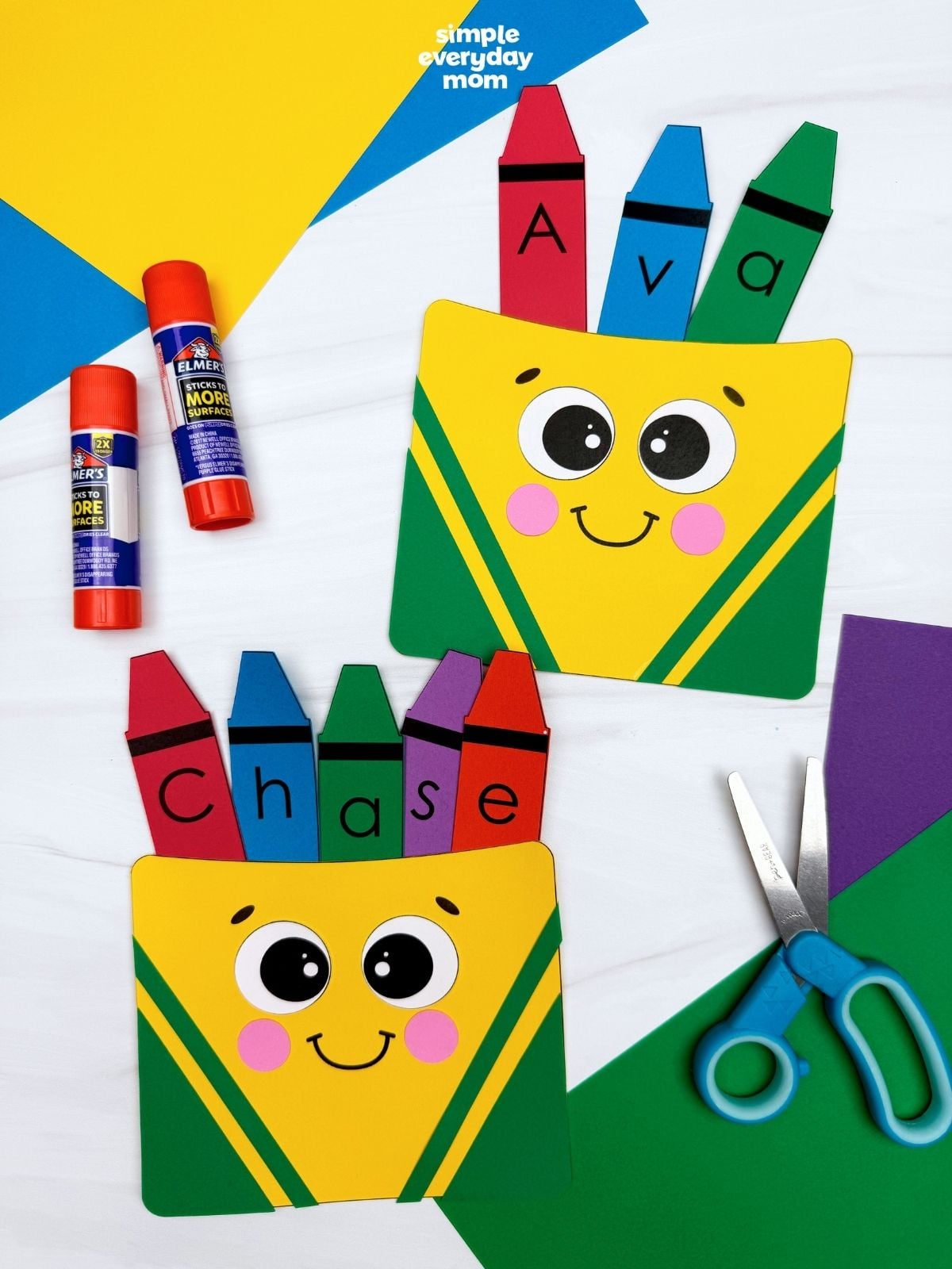 Two finished personalized crayon box crafts displayed with glue sticks, scissors, and colored paper.