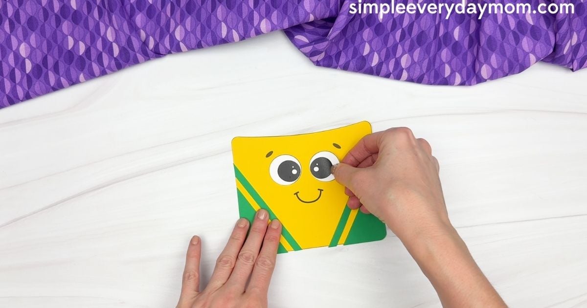 Hand placing the second eye on the smiling crayon box craft.