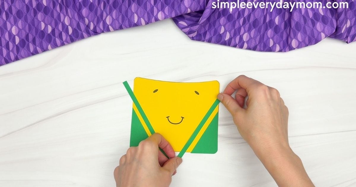 Hands attaching narrow green strips to the front of the crayon box craft.
