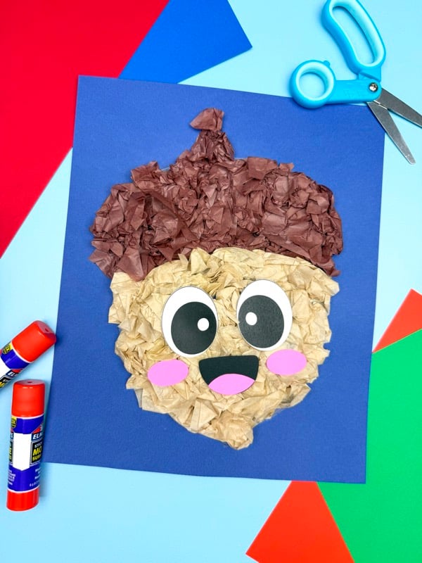 Acorn tissue paper craft on blue background with glue sticks, scissors, and colorful paper for a fun fall activity.